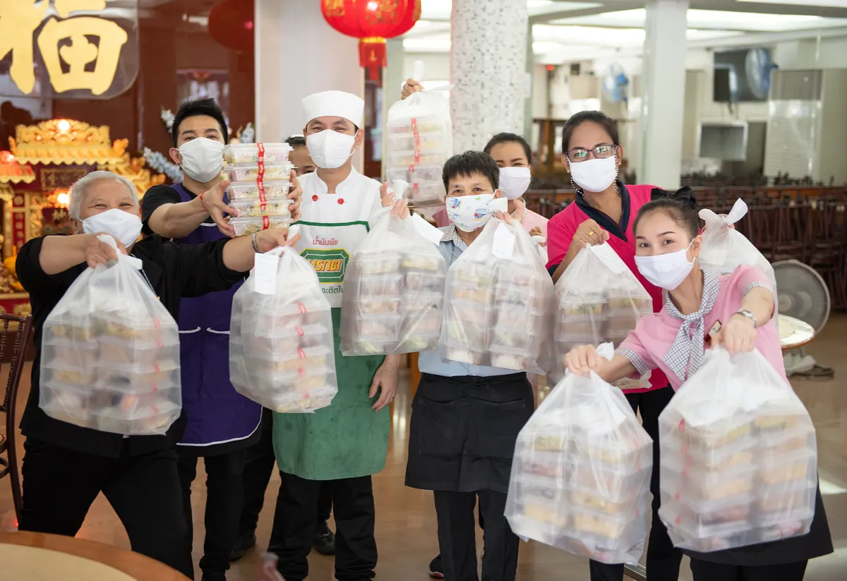 ทีมพนักงานและเชฟร้าน Ruenpetch Suki สวมหน้ากากอนามัย ยิ้มแย้มพร้อมให้บริการ โดยถือถุงบรรจุกล่องอาหารเดลิเวอรี่จำนวนมากเตรียมจัดส่งให้ลูกค้า โดยมีฉากหลังเป็นบรรยากาศภายในร้านและศาลเจ้าจีนสไตล์ดั้งเดิม